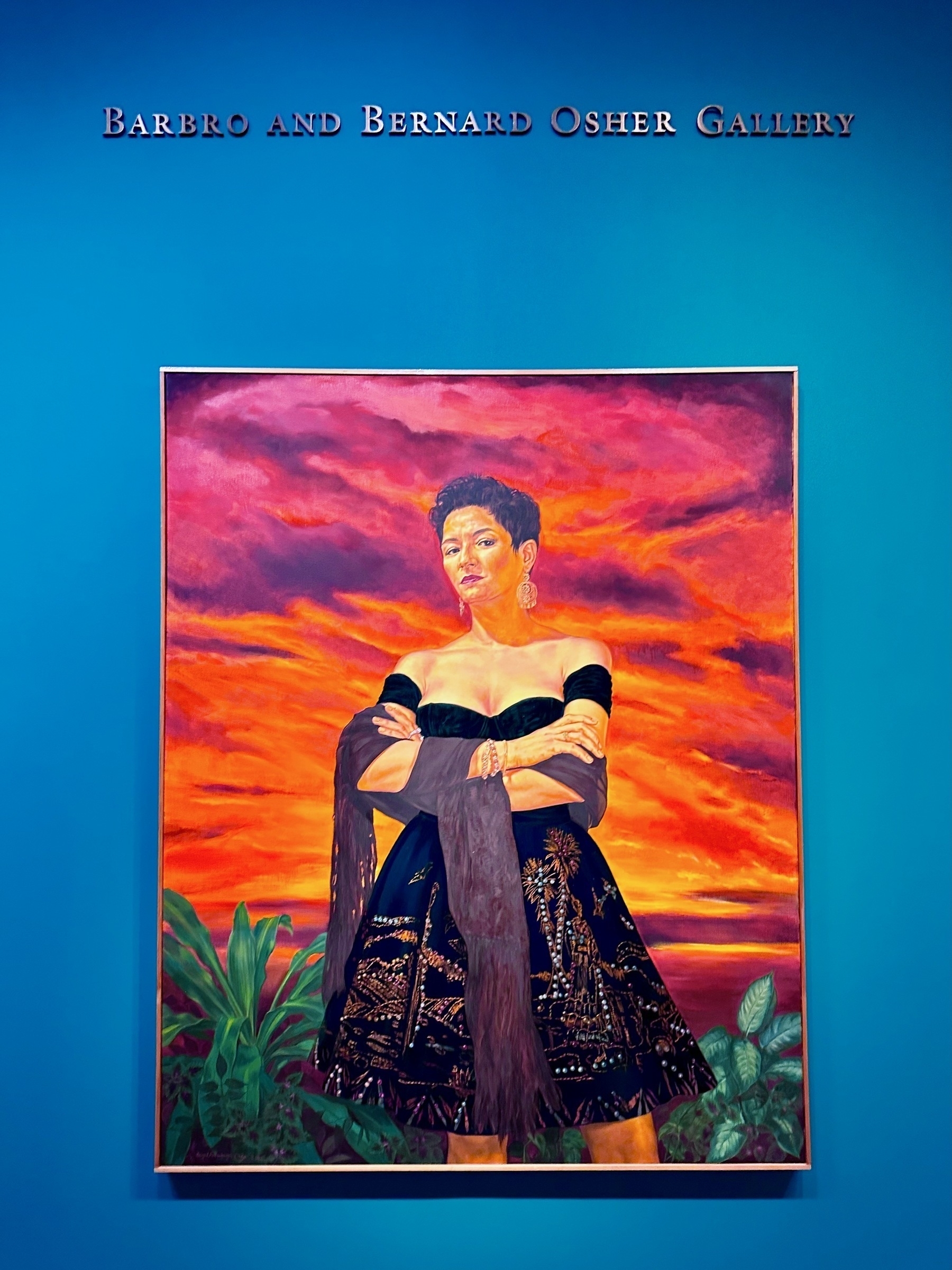 Portrait of a woman standing in front of a purple-red sunset in a black gown, arms crossed, looking straight at you.