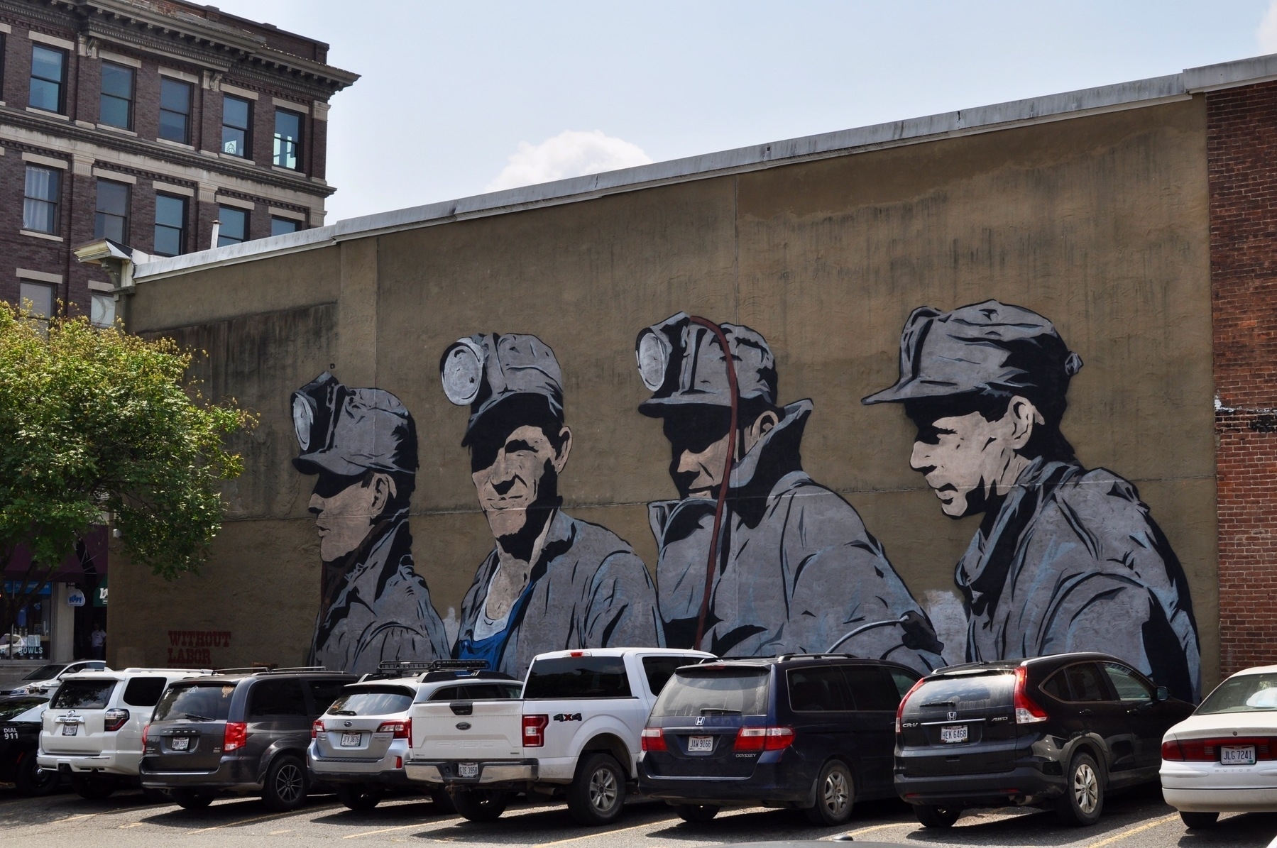 Photo of a mural of four coal miners in gray uniforms. In front of the mural is a row of parked SUVs, mini vans, and pickup trucks.