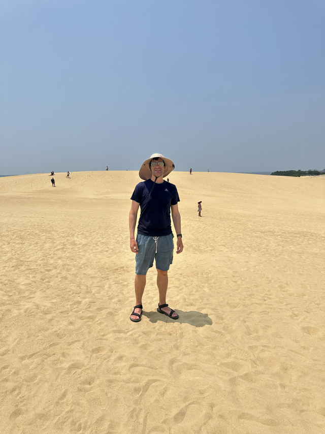 Photo of the author standing on a sand dune wearing a large sun-blocking hat and sunglassess. Several tiny figures are in the background.