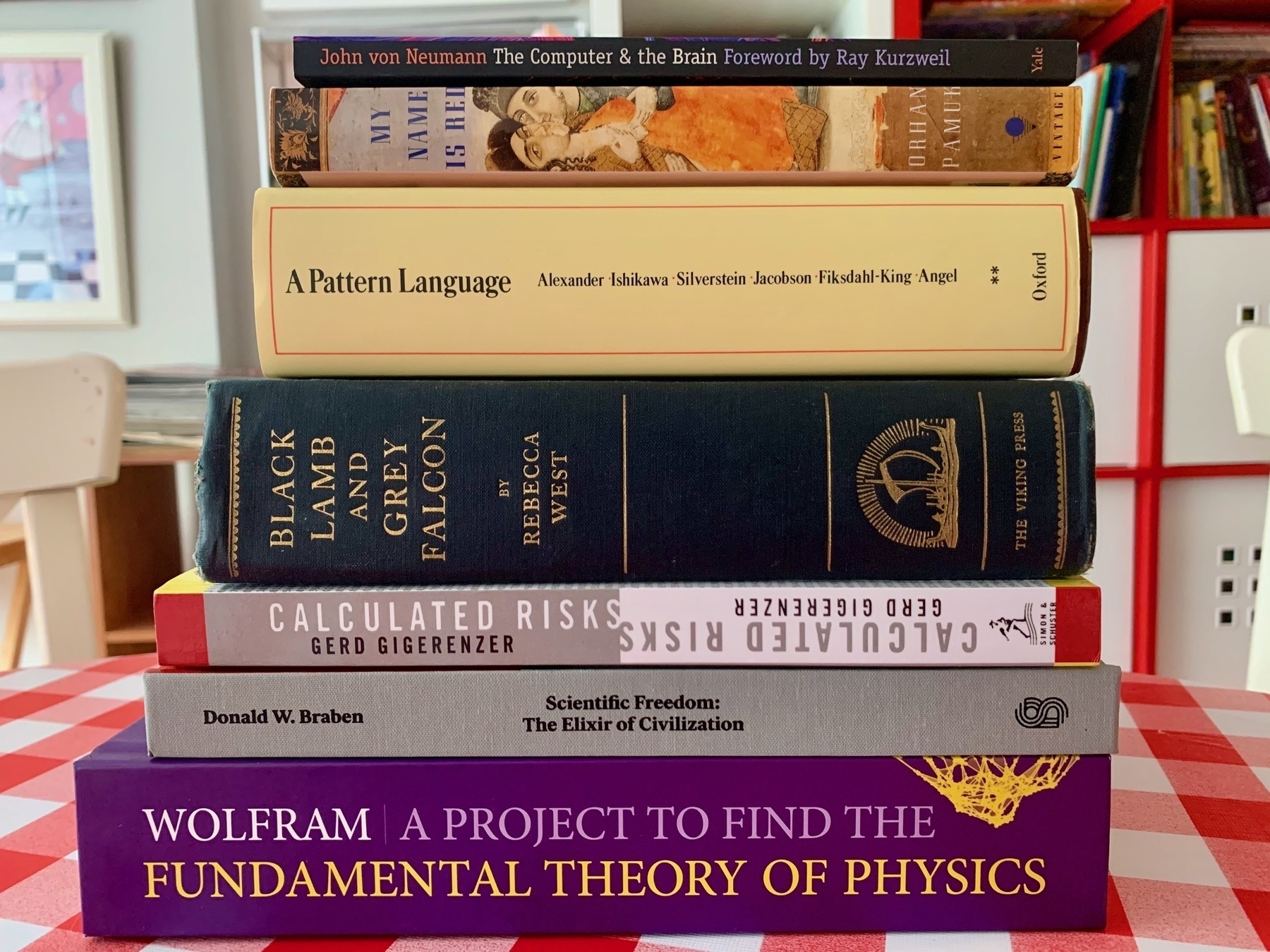 A stack of seven books laying on a kitchen table with only spines showing. The thickest are &quot;Black Lamb and Gray Falcon&quot; and &quot;A Pattern Language&quot;. Wolfram&#39;s &quot;Fundamental Theory of Physics&quot; is at the base.