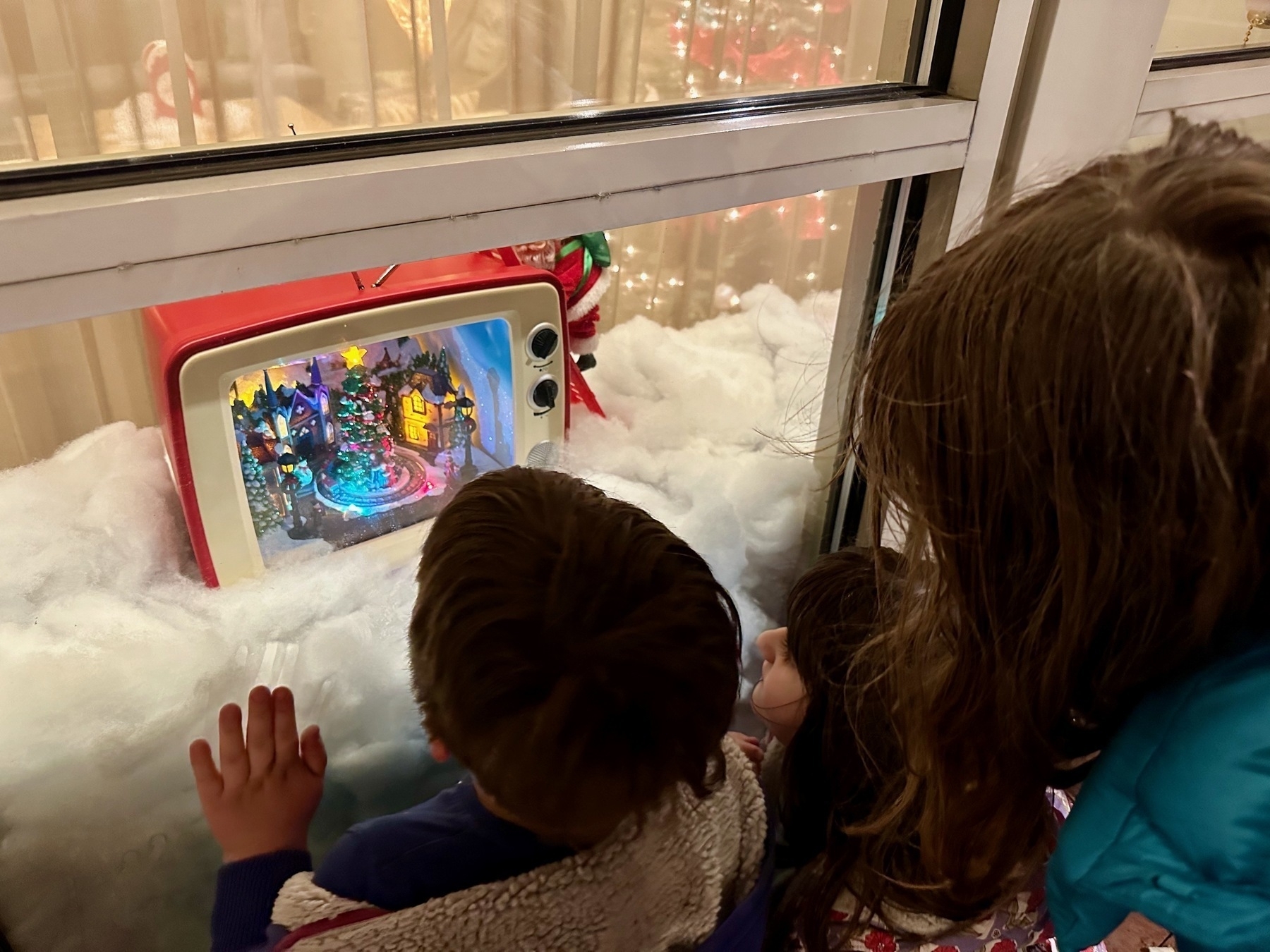 Children looking through a shopping window at a holiday display featuring an old toy TV set with a decorated Christmas tree inside.