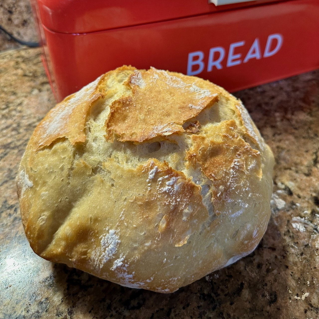 A rustic loaf of crusty bread in front of a red bred box.