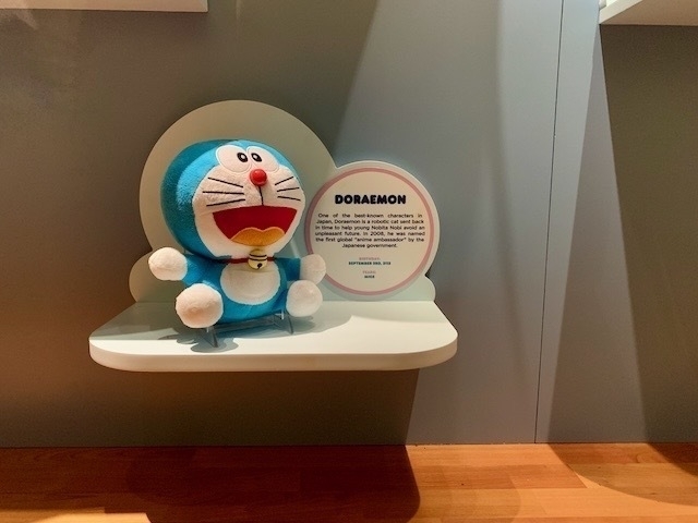 Doraemon plush toy in a museum case. Text next to it lists a September 3rd, 2112 birthday.