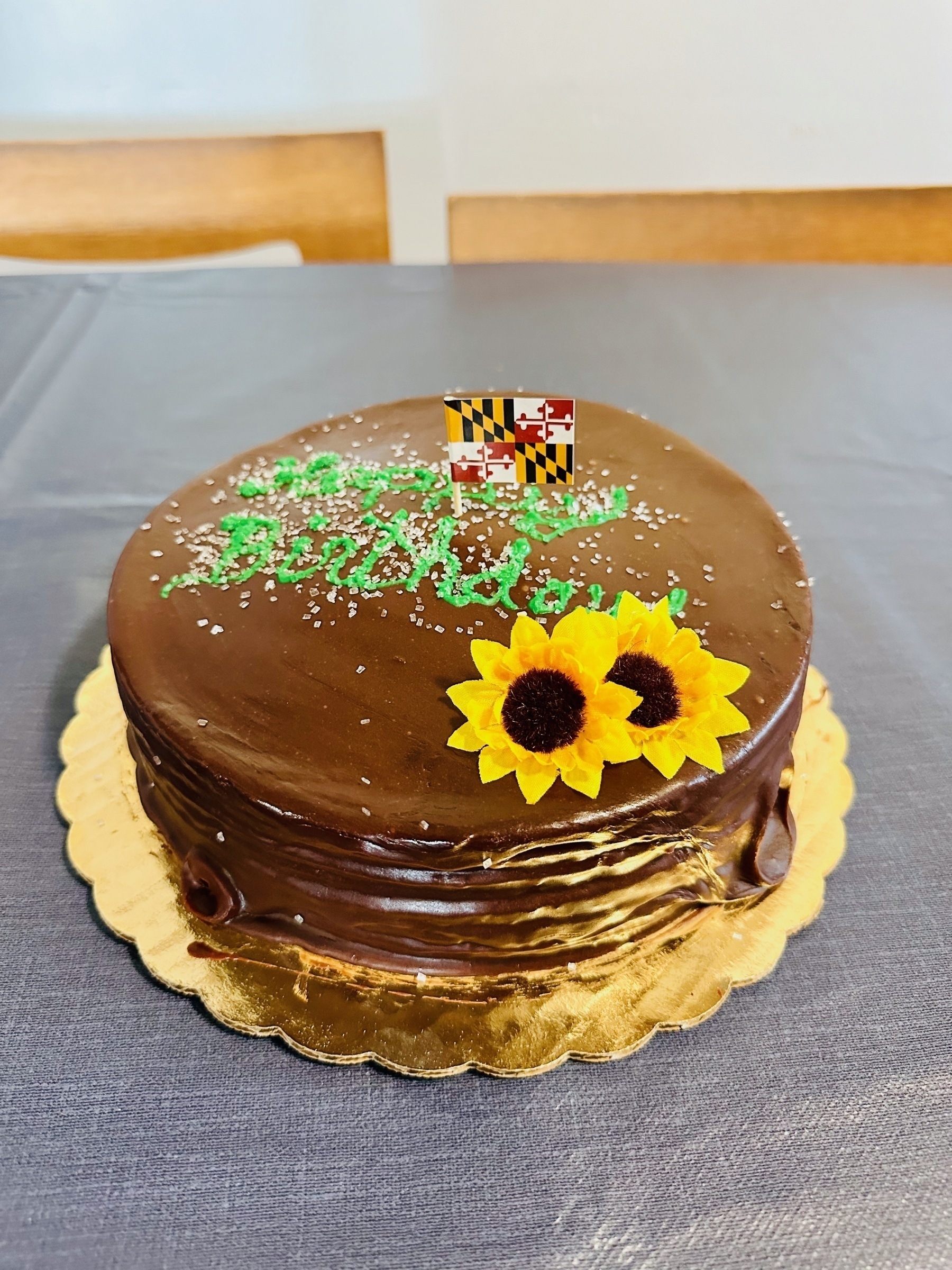 A birthday cake with chocolate glazing, two (plastic) black-eyed Susans and a (paper) Maryland state flag placed on top.