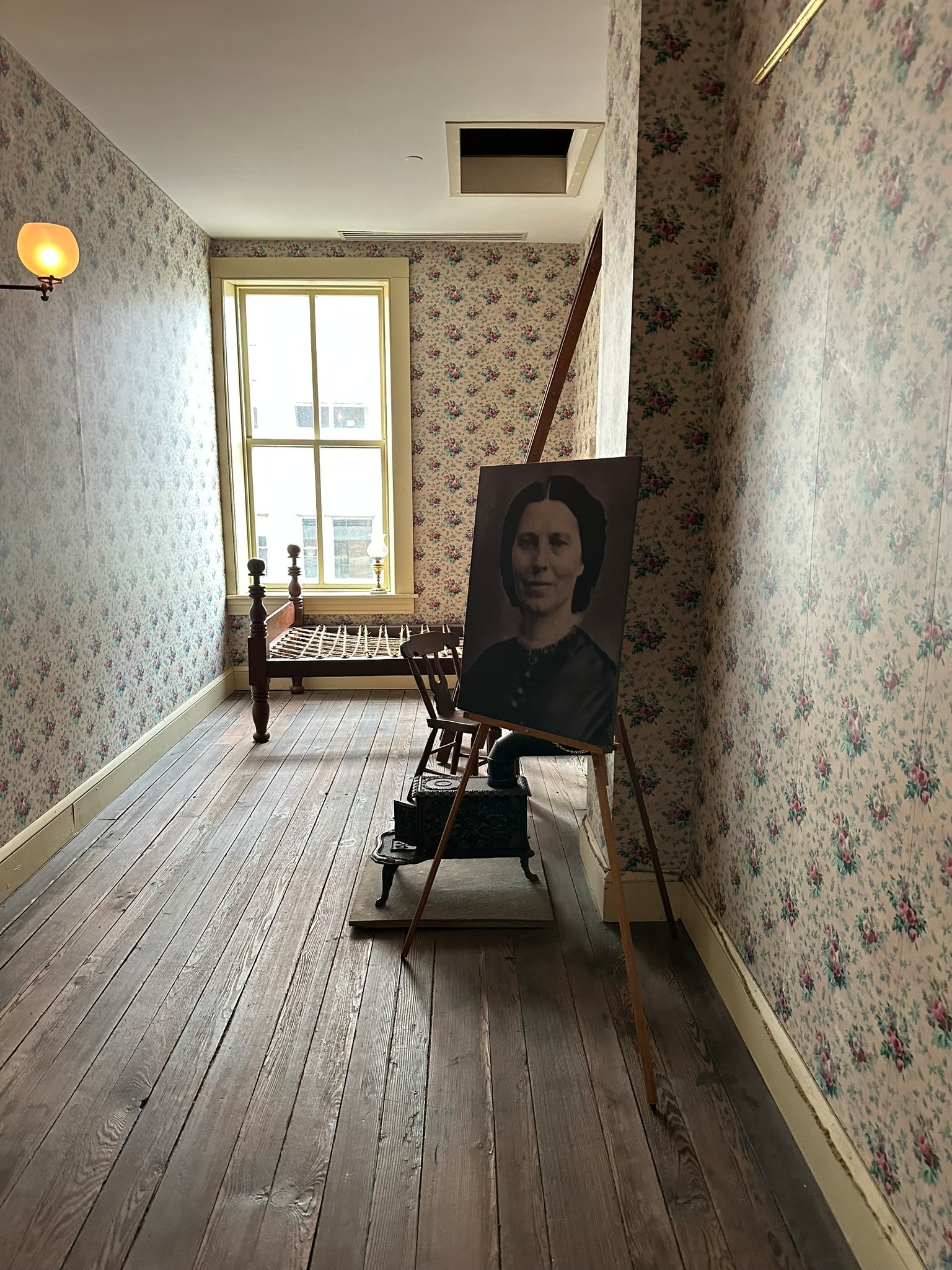 A small bedroom, as wide as a twin bed in it is long, containing a bed, an iron furnace, and a photo of Clara Barton.